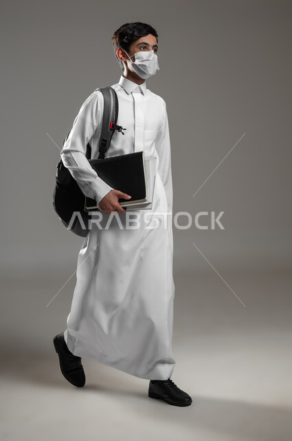 ارتداء الكمامة للوقاية من الأمراض، المشي والذهاب الى المدرسة، متابعة الدروس والواجبات اليومية، بورتريه لولد عربي سعودي خليجي يرتدي الثوب التقليدي وحقيبة الظهر يستعد لبداية العام الدراسي الجديد, صورة الجسم بالكامل، خلفية رمادية، محتوى حساس، استخدام حساس