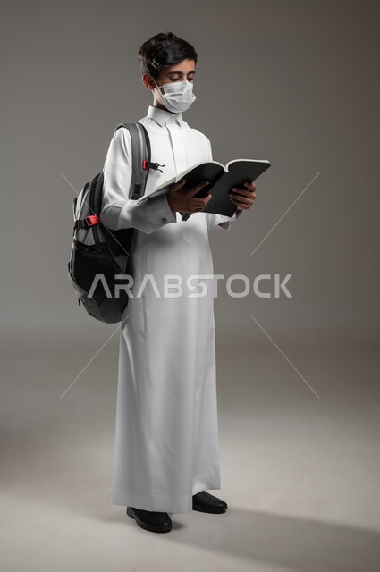 اتباع الاجراءات الوقائية، الاجتهاد وتحضير الدروس في بداية العام الدراسي، متابعة الواجبات اليومية، بورتريه لولد عربي سعودي خليجي يرتدي الثوب التقليدي يحمل الحقيبة المدرسية يضع الكمامة يقوم بقراءة الكتب, صورة الجسم بالكامل، خلفية رمادية، محتوى حساس، استخدام حساس