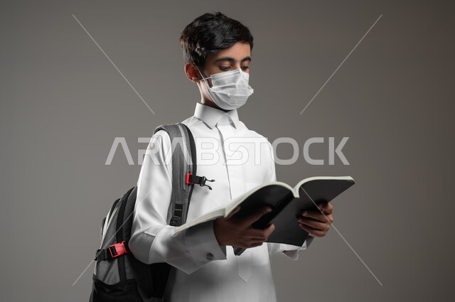 اتباع الاجراءات الوقائية، الاجتهاد وتحضير الدروس في بداية العام الدراسي الجديد, متابعة الدروس والواجبات اليومية، بورتريه مقرب لولد عربي سعودي خليجي يرتدي الثوب التقليدي يحمل الحقيبة المدرسية يضع الكمامة يقوم بقراءة الكتب, خلفية رمادية، محتوى حساس، استخدام حساس