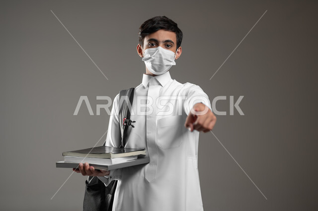 النظر إلى الكاميرا والإشارة الى شيء ما، الالتزام بالإجراءات الوقائية، متابعة الدروس والواجبات، بورتريه مقرب لولد عربي سعودي خليجي يرتدي الثوب التقليدي وحقيبة الظهر يضع الكمامة يمسك كتبه المدرسية، خلفية رمادية، محتوى حساس، استخدام حساس