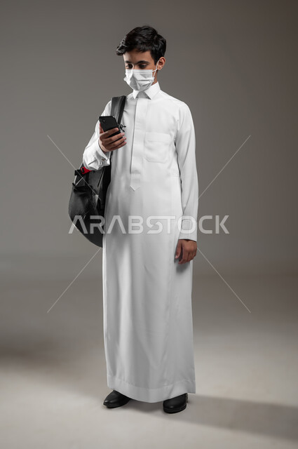 Following up on assignments online via mobile phone, adhering to preventive measures, integrating science with technology and technology, a portrait of a Saudi Arabian Gulf student wearing a traditional dress and a backpack, wearing a mask, using a mobile phone, a full-body photo, gray background, sensitive use, sensitive content.