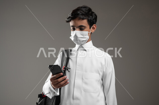 الالتزام بالإجراءات الوقائية، دمج العلم مع التقنية والتكنوجيا، متابعة الواجبات اونلاين عبر الهاتف المحمول، بورتريه مقرب لطالب عربي خليجي سعودي يرتدي الثوب التقليدي وحقيبة الظهر يضع الكمامة يقوم باستعمال الموبايل، خلفية رمادية، محتوى حساس، استخدام حساس