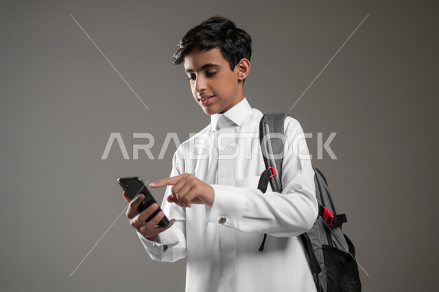 تطور مجال التعلم في السعودية، استعمال الأجهزة التقنية والإلكترونية الحديثة، متابعة الواجبات اونلاين عبر الهاتف المحمول، بورتريه مقرب لطالب عربي خليجي سعودي مبتسم يرتدي الثوب التقليدي وحقيبة الظهر ينقر على شاشة الموبايل، خلفية رمادية، محتوى حساس، استخدام حساس