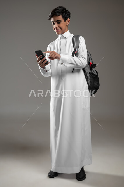 متابعة الواجبات اونلاين، استعمال الأجهزة الإلكترونية الحديثة بالتعلم، دمج العلم مع التقنية والتكنوجيا، بورتريه لطالب عربي خليجي سعودي مبتسم يرتدي الثوب التقليدي وحقيبة الظهر ينقر على شاشة الموبايل، صورة الجسم بالكامل، خلفية رمادية، استخدام حساس، محتوى حساس