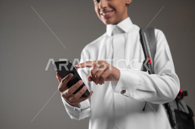 Using modern electronic devices for learning, integrating science with technology and technology, following up on assignments online via mobile phone, close-up portrait of a smiling Saudi Arabian Gulf student wearing a traditional dress and backpack, tapping on the mobile screen, gray background, sensitive content, sensitive use