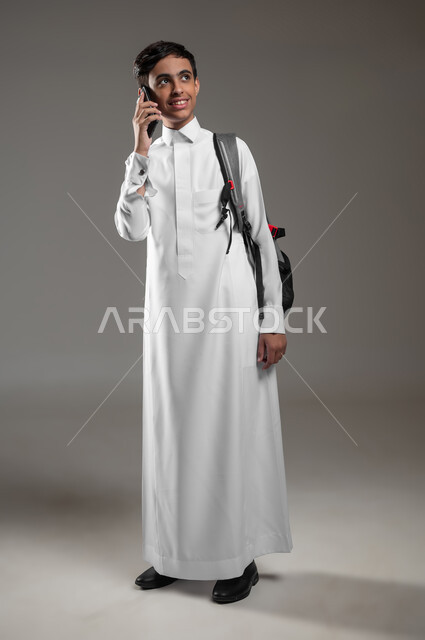 النظر الى شيء ما بتعابير السرور والسعادة، بورتريه لطالب عربي خليجي سعودي مبتسم يرتدي الثوب التقليدي وحقيبة الظهر يقوم بالرد والإجابة على اتصال هاتفي، التواصل والدردشة مع الأصدقاء، صورة الجسم بالطول الكامل، خلفية رمادية، محتوى حساس، استخدام حساس