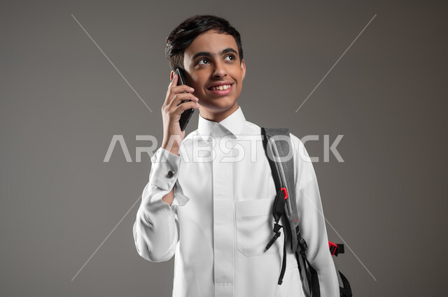 التواصل والدردشة مع الأصدقاء والأقارب باستعمال الموبايل، النظر الى شيء ما بتعابير السرور والسعادة، بورتريه مقرب لطالب عربي خليجي سعودي مبتسم يرتدي الثوب التقليدي وحقيبة الظهر يقوم بالرد والإجابة على اتصال هاتفي، خلفية رمادية، محتوى حساس، استخدام حساس