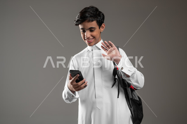 إجراء مكالمات اونلاين باستعمال الهاتف المحمول، رفع اليد بايماءات إلقاء التحية، بورتريه مقرب لطالب عربي خليجي سعودي مبتسم يرتدي الثوب التقليدي وحقيبة الظهر يقوم بالدردشة مع الأصدقاء والأقارب بتعابير السرور والسعادة، خلفية رمادية، محتوى حساس، استخدام حساس