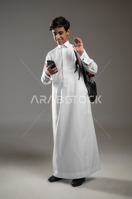 رفع اليد بايماءات إلقاء التحية، التواصل والدردشة مع الأصدقاء بتعابير السرور والسعادة، بورتريه لطالب عربي خليجي سعودي يرتدي الثوب التقليدي وحقيبة الظهر يقوم بإجراء مكالمات اونلاين باستعمال الهاتف المحمول، صورة لكامل الجسم، خلفية رمادية، محتوى حساس، استخدام حساس