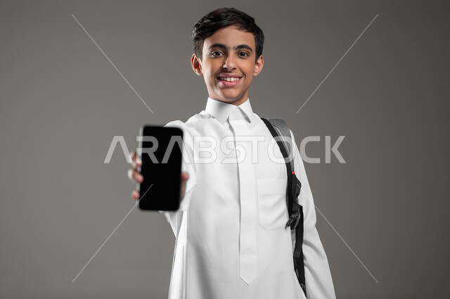 عرض هاتف محمول بشاشة سوداء فارغة، دمج العلم بالتكنولوجيا، استخدام الأجهزة الالكترونية الحديثة في مجال التعليم، بورتريه مقرب لطالب عربي خليجي سعودي يرتدي الثوب التقليدي وحقيبة الظهر ينظر الى الكاميرا بتعابير السرور والفرح، خلفية رمادية، محتوى حساس، استخدام حساس