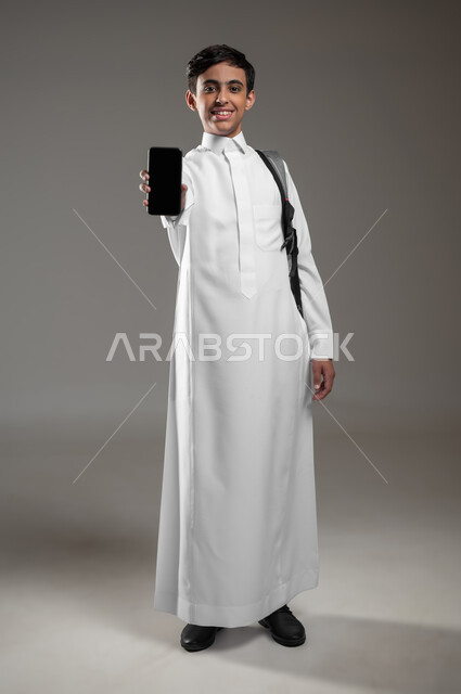 دمج العلم بالتكنولوجيا، استخدام الأجهزة الالكترونية الحديثة في مجال التعليم، بورتريه لطالب عربي خليجي سعودي مبتسم يرتدي الثوب التقليدي وحقيبة الظهر ينظر الى الكاميرا، عرض هاتف محمول بشاشة سوداء فارغة، صورة لكامل الجسم، خلفية رمادية، محتوى حساس، استخدام حساس