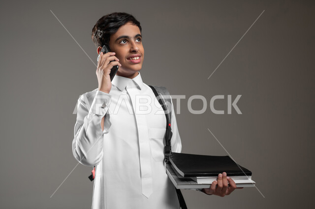 إجراء مكالمة عبر الهاتف المحمول، التواصل والدردشة مع الأصدقاء، التعليم في المملكة السعودية، النظر الى شيء ما بتعابير الفرح، بورتريه مقرب لطالب عربي خليجي سعودي مبتسم يرتدي الثوب التقليدي وحقيبة الظهر يحمل الكتب الدراسية، خلفية رمادية، محتوى حساس، استخدام حساس