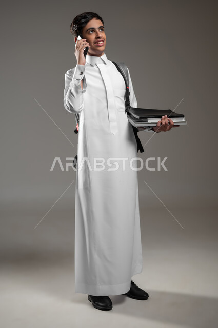 النظر الى شيء ما بتعابير السرور، بورتريه لطالب عربي خليجي سعودي مبتسم يرتدي الثوب التقليدي وحقيبة الظهر يحمل الكتب الدراسية، إجراء مكالمات عبر الهاتف المحمول، التواصل والدردشة مع الأصدقاء، صورة لكامل الجسم، خلفية رمادية، محتوى حساس، استخدام حساس