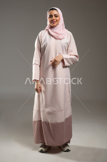 The concept of femininity and elegance, concern for external appearance, gestures of pleasure and happiness, portrait of a smiling Saudi Arabian Gulf woman wearing a pink abaya looking at the camera with expressions of joy, full-length body image, gray background, sensitive content, sensitive use
