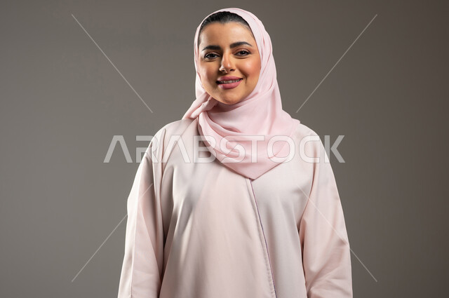 الاهتمام بالمظهر الخارجي، مفهوم الانوثة والاناقة، ايماءات السرور والسعادة، بورتريه مقرب لامرأه عربيه خليجيه سعوديه محجبة مبتسمة ترتدي عباءة وردية تنظر إلى الكاميرا بتعابير الفرح، خلفية رمادية، محتوى حساس، استخدام حساس