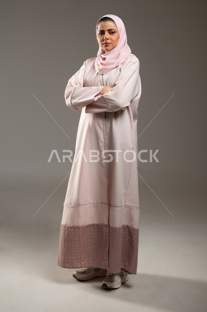 النظر إلى الكاميرا بايماءات الحزم والجدية، تعابير الثقة بالنفس، الاهتمام بالمظهر الخارجي، بورتريه لامرأة عربية خليجية سعودية محجبة ترتدي عباءة وردية تقف بأيدي متقاطعة، صورة الجسم بالطول الكامل، خلفية رمادية، محتوى حساس، استخدام حساس