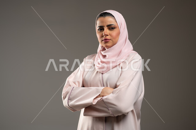 ايماءات الحزم والجدية، الاهتمام بالمظهر الخارجي، تعابير الثقة بالنفس، بورتريه مقرب لامرأة عربية خليجية سعودية محجبة ترتدي عباءة وردية تقف بأيدي متقاطعة، خلفية رمادية، محتوى حساس، استخدام حساس