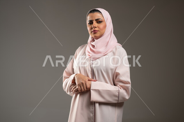 النظر إلى الكاميرا بايماءات الحزم والجدية، تعابير الثقة بالنفس، الاهتمام بالمظهر الخارجي، بورتريه مقرب لامرأة عربية خليجية سعودية محجبة ترتدي عباءة وردية تقف بأيدي متقاطعة، خلفية رمادية، محتوى حساس، استخدام حساس