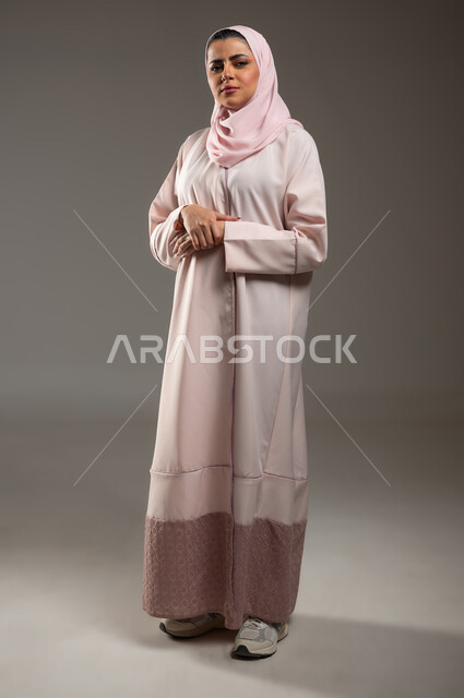 تعابير الثقة بالنفس، النظر إلى الكاميرا بايماءات الحزم والجدية، الاهتمام بالمظهر الخارجي، بورتريه لامرأة عربية خليجية سعودية محجبة ترتدي عباءة وردية تقف بأيدي متقاطعة، صورة الجسم بالطول الكامل، خلفية رمادية، محتوى حساس، استخدام حساس