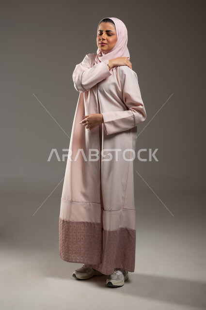 التهاب العضلات والمفاصل، الحاجه للعلاج وطلب المساعده، الإحساس بالمرض والتعب، بورتريه من الجانب لامرأه عربيه خليجيه سعوديه ترتدي عباءة وردية تضع يدها على كتفها بإيماءات تدل على الألم، صورة الجسم بالطول الكامل، خلفية رمادية، محتوى حساس، استخدام حساس