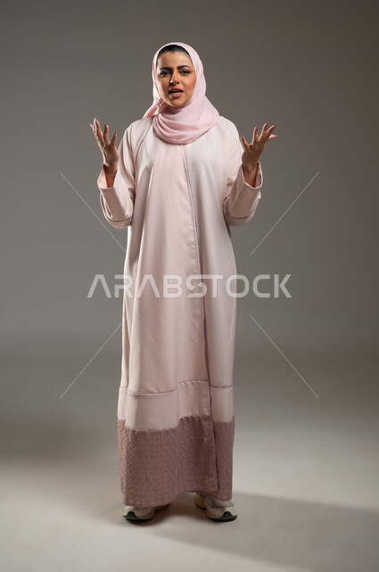 الشعور بالانزعاج والقهر، بورتريه لامرأه عربيه خليجيه سعودية محجبة ترتدي عباءة وردية ترفع يديها بإيماءات وجه ويدين تدل على الانفعال الشديد،  مفهوم الصراخ والغضب، صورة الجسم بالطول الكامل، خلفية رمادية، محتوى حساس، استخدام حساس
