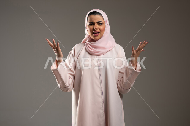 The concept of screaming and anger, a close-up portrait of a veiled Saudi Arabian Gulf woman wearing a pink abaya, raising her hands with facial and hand gestures indicating extreme emotion, feeling of annoyance and oppression, gray background, sensitive content, sensitive use
