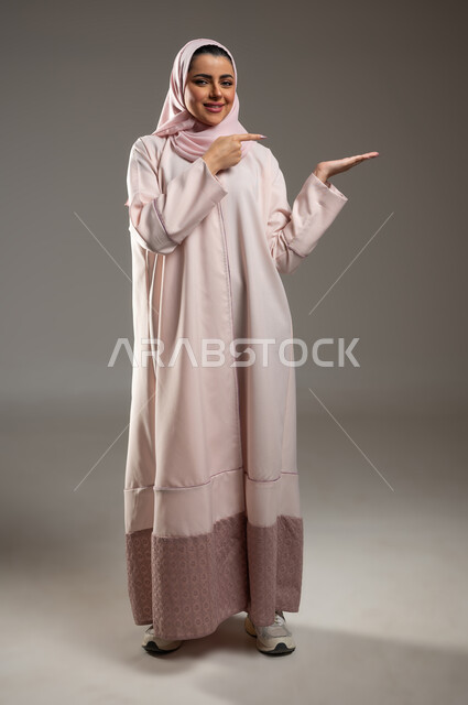 النظر إلى الكاميرا وتحديد شيء معين بإيماءات السرور, بورتريه لامرأة عربية خليجية سعودية محجبة مبتسمة ترتدي الثوب عباءة وردية تفتح كفها للأعلى وتشير إلى عنصر ما، صورة الجسم بالطول الكامل, خلفية رمادية، محتوى حساس، استخدام حساس