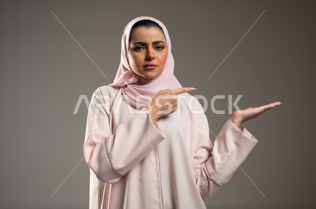 تعابير الحزم والجدية، النظر إلى الكاميرا وتحديد شيء معين بإيماءات الثقة بالنفس, بورتريه مقرب لامرأة عربية خليجية سعودية محجبة ترتدي الثوب عباءة وردية تفتح كفها للأعلى وتشير إلى عنصر ما، خلفية رمادية، محتوى حساس، استخدام حساس