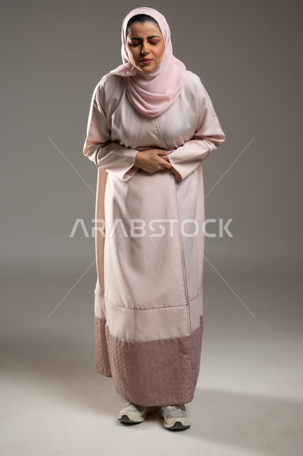 الشعور بحرقة في المعدة وعسر الهضم، الحاجه للمساعدة وطلب العلاج، الإحساس بالمغص والوجع، بورتريه لامرأه عربيه خليجيه سعوديه ترتدي عباءة وردية تضع يدها على بطنها تنحني من شدة الألم، إيماءات التقيؤ والغثيان، صورة لكامل الجسم، خلفية رمادية، محتوى حساس، استخدام حساس