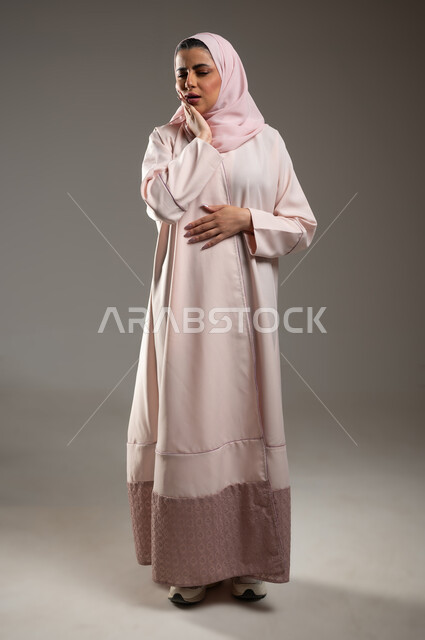 الحاجه لزيارة الطبيب، طلب العلاج و المساعده، تسوس الأضراس وإلتهاب اللثه، بورتريه لامرأه عربيه خليجيه سعوديه محجبة ترتدي عباءة وردية تضع يدها على خدها بإيماءات تدل على الشعور بألم الأسنان، صورة الجسم بالطول الكامل، خلفية رمادية، محتوى حساس، استخدام حساس