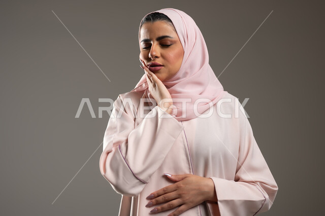 طلب العلاج و المساعده، تسوس الأضراس وإلتهاب اللثه، بورتريه مقرب لامرأه عربيه خليجيه سعوديه محجبة ترتدي عباءة وردية تضع يدها على خدها بإيماءات تدل على الشعور بألم الأسنان، الحاجه لزيارة الطبيب، خلفية رمادية، محتوى حساس، استخدام حساس