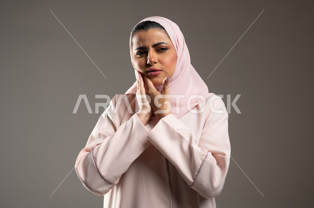 Seeking treatment and assistance, tooth decay and gingivitis, close-up portrait of a veiled Saudi Arabian Gulf woman wearing a pink abaya, placing her hand on her cheek with gestures indicating a feeling of toothache, the need to visit a doctor, gray background, sensitive content, sensitive use