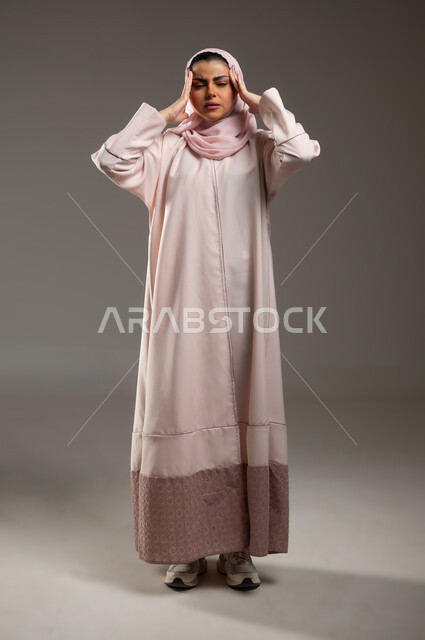 الإحساس بألم الرأس والصداع، المعاناة من آلام الشقيقة، الحاجة للهدوء والراحة، بورتريه لإمرأة عربية خليجية سعودية محجبة ترتدي عباءة وردية تضع يديها على رأسها بايماءات التعب والارهاق، صورة الجسم بالطول الكامل، خلفية رمادية، محتوى حساس، استخدام حساس