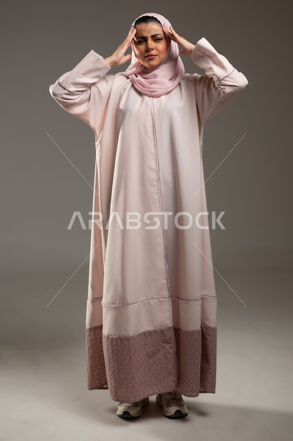 الحاجة للهدوء والراحة، الإحساس بألم الرأس والصداع، المعاناة من آلام الشقيقة، بورتريه لإمرأة عربية خليجية سعودية محجبة ترتدي عباءة وردية تضع يديها على رأسها بايماءات التعب والارهاق، صورة الجسم بالطول الكامل، خلفية رمادية، محتوى حساس، استخدام حساس