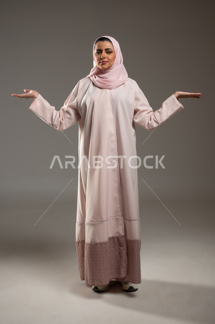 Feeling confused and ignorant about something, hesitation and lack of knowledge about a certain thing, portrait of a veiled Saudi Arabian Gulf woman wearing a pink abaya raising her hands with gestures of uncertainty, full-length body image, gray background, sensitive content, sensitive use