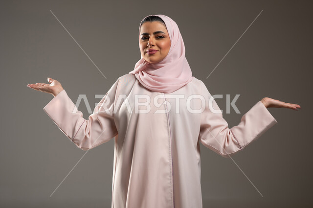 التردد وانعدام المعرفة بخصوص شيء معين، الشعور بالحيرة والجهل بأمر ما، بورتريه مقرب لامرأة عربية خليجية سعودية محجبة ترتدي عباءة وردية ترفع يديها بإيماءات عدم التأكد، خلفية رمادية، محتوى حساس، استخدام حساس