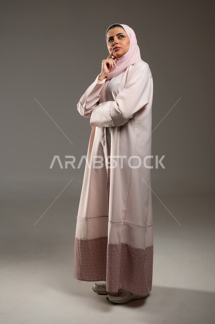 التمعن والتركيز والتخطيط للمستقبل، محاولة إيجاد حل لمشكلة ما، بورتريه لإمرأة عربية خليجية سعودية محجبة ترتدي عباءة وردية تضع يدها على ذقنها وتنظر إلى الأعلى بإيماءات التفكير والتأمل، صورة الجسم بالطول الكامل، خلفية رمادية، محتوى حساس، استخدام حساس