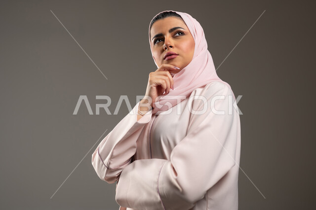 Trying to find a solution to a problem, contemplating, concentrating and planning for the future, a close-up portrait of a veiled Saudi Arabian Gulf woman wearing a pink abaya, placing her hand on her chin and looking up with gestures of thought and contemplation, gray background, sensitive content, sensitive use