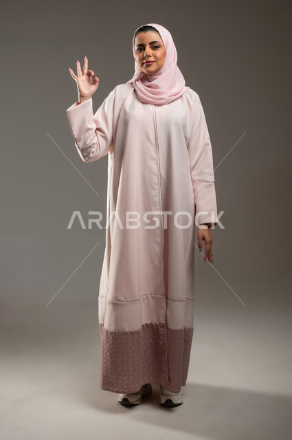 مفهوم القبول والموافقه، علامة الجوده والإمتياز، بورتريه لامرأه عربيه خليجيه سعوديه ترتدي عباءة وردية ترفع يدها بإيماءات تدل على الإعجاب والرضى، صورة الجسم بالطول الكامل، خلفية رمادية، محتوى حساس، استخدام حساس