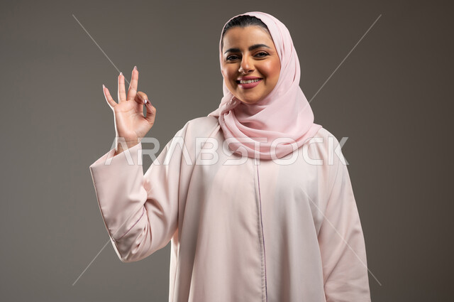تعابير السرور والسعادة، علامة الجوده والإمتياز، مفهوم القبول والموافقه، بورتريه مقرب لامرأه عربيه خليجيه سعوديه ترتدي عباءة وردية ترفع يدها بإيماءات تدل على الإعجاب والرضى، خلفية رمادية، محتوى حساس، استخدام حساس