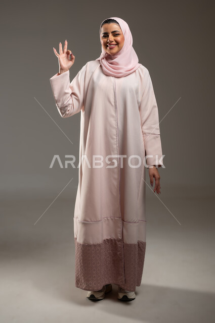 النظر إلى الكاميرا بتعابير السرور والسعادة، علامة الجوده والإمتياز، مفهوم القبول والموافقه، بورتريه لامرأه عربيه خليجيه سعوديه ترتدي عباءة وردية ترفع يدها بإيماءات تدل على الإعجاب والرضى، صورة الجسم بالطول الكامل، خلفية رمادية، محتوى حساس، استخدام حساس