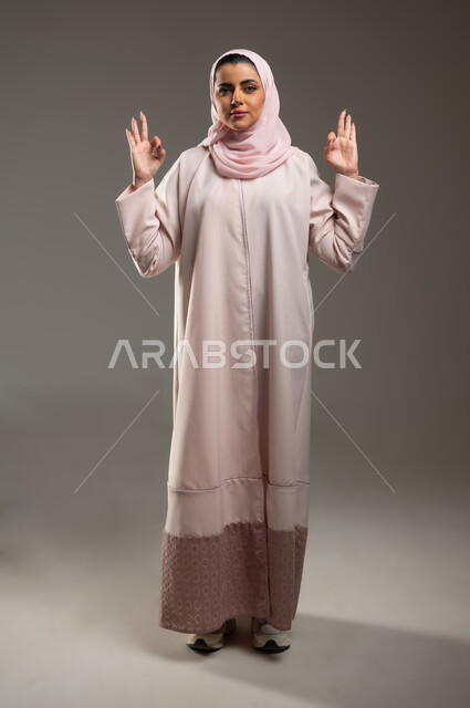 مفهوم القبول والموافقه، علامة الجوده والإمتياز، بورتريه لامرأه عربيه خليجيه سعوديه ترتدي عباءة وردية ترفع يديها بإيماءات تدل على الإعجاب والرضى، صورة الجسم بالطول الكامل، خلفية رمادية، محتوى حساس، استخدام حساس