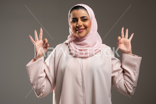 تعابير السرور والسعادة، علامة الجوده والإمتياز، بورتريه مقرب لامرأه عربيه خليجيه سعوديه ترتدي عباءة وردية ترفع يديها بإيماءات تدل على الإعجاب والرضى، مفهوم القبول والموافقه، خلفية رمادية، محتوى حساس، استخدام حساس