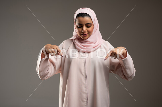 إشارات وحركات يدين تدل على هذا المكان (هنا)، تحديد موقع شيء معين، بورتريه مقرب لامرأة عربية خليجية سعودية محجبة ترتدي عباءة وردية تشير بإصبعي السبابة إلى جهة الأسفل، خلفية رمادية، محتوى حساس، استخدام حساس