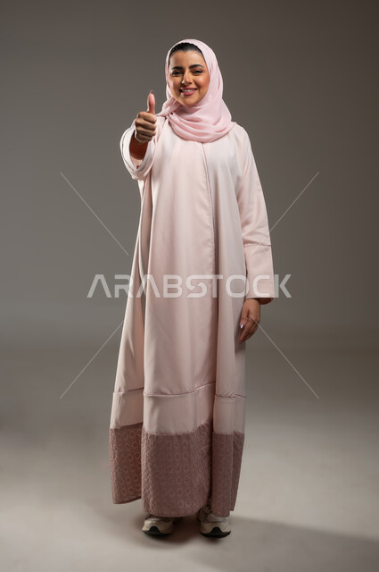 النظر إلى الكاميرا بتعابير الإعجاب والرضى، مفهوم القبول والموافقه، بورتريه لامرأه عربيه خليجيه سعوديه محجبة ترتدي عباءة وردية ترفع اصبع الابهام بإيماءات الامتياز والجودة، صورة الجسم بالطول الكامل، خلفية رمادية، محتوى حساس، استخدام حساس