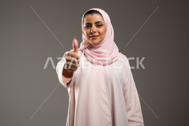 مفهوم القبول والموافقه، تعابير الإعجاب والرضى، بورتريه مقرب لامرأه عربيه خليجيه سعوديه محجبة ترتدي عباءة وردية ترفع اصبع الابهام بإيماءات الامتياز والجودة، خلفية رمادية، محتوى حساس، استخدام حساس