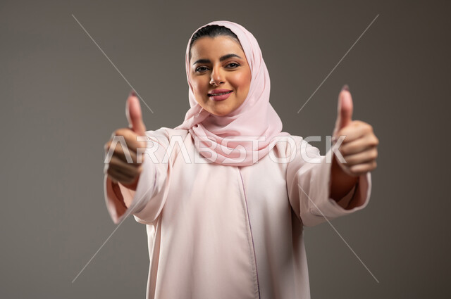 تعابير الإعجاب والرضى، بورتريه مقرب لامرأه عربيه خليجيه سعوديه محجبة ترتدي عباءة وردية ترفع اصبع الابهام بإيماءات الامتياز والجودة، مفهوم القبول والموافقه، خلفية رمادية، محتوى حساس، استخدام حساس