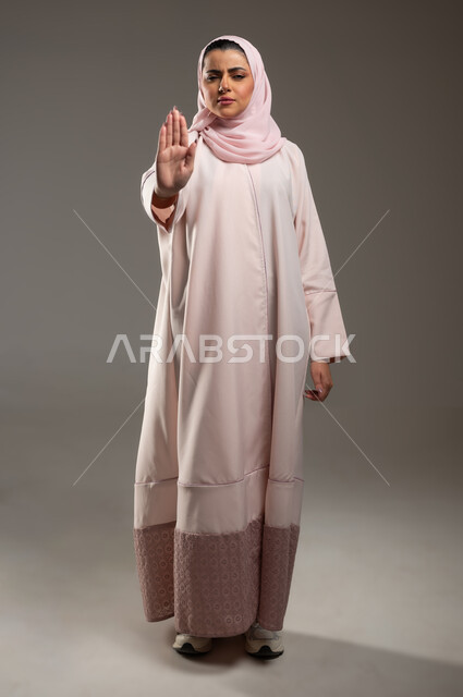 Feelings of annoyance and anger, expressions of rejection and abstention, the command to stop doing something, a portrait of a Saudi Gulf Arab woman, Hajjah, wearing a pink abaya, raising her hand with gestures indicating prohibition, full-length body image, gray background, sensitive content, sensitive use