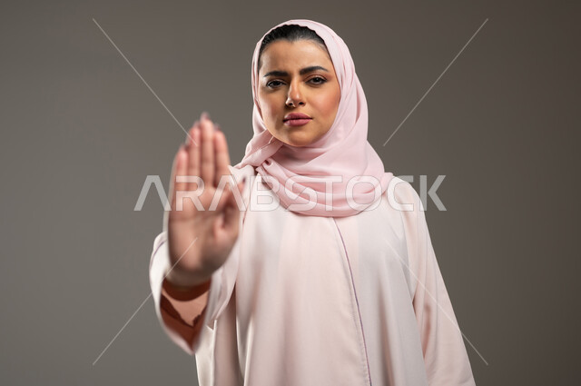 تعابير الرفض والإمتناع، الأمر بالتوقف عن فعل شيء ما، بورتريه مقرب لامرأه عربيه خليجيه سعوديه محجة ترتدي عباءة وردية ترفع يدها بإيماءات تدل على النهي، الشعور بالإنزعاج والغضب، خلفية رمادية، محتوى حساس، استخدام حساس