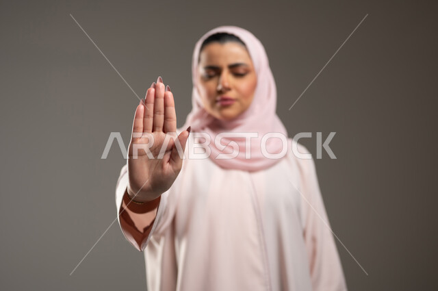 The command to stop doing something, a close-up portrait of a Saudi Gulf Arab woman, a woman wearing a pink abaya, raising her hand with gestures indicating prohibition, feeling annoyed and angry, expressions of rejection and abstention, gray background, sensitive content, sensitive use